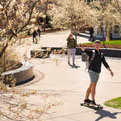 male student on a long board crossing campus 